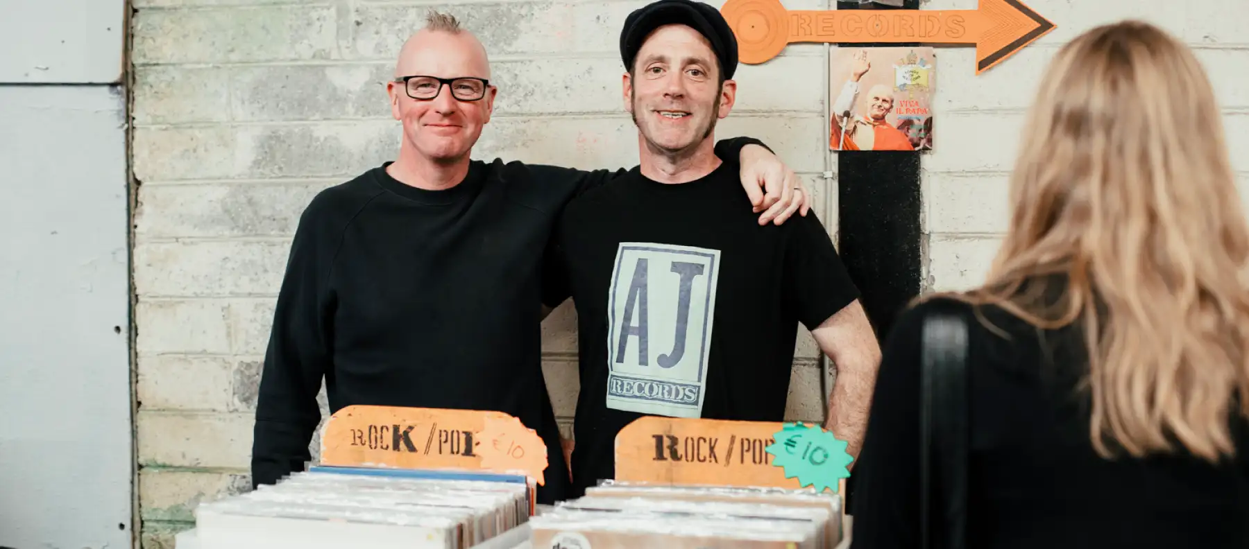 Two men smiling and standing in front of a vintage vinyl stall at a market